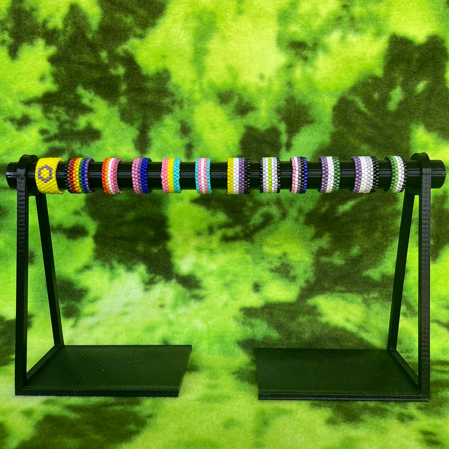 A set of beaded pride rings on a black display set on a light green and dark green tie dye backdrop. there are 12 rings, each reprisenting a different pride flag. this rings are thick in width.