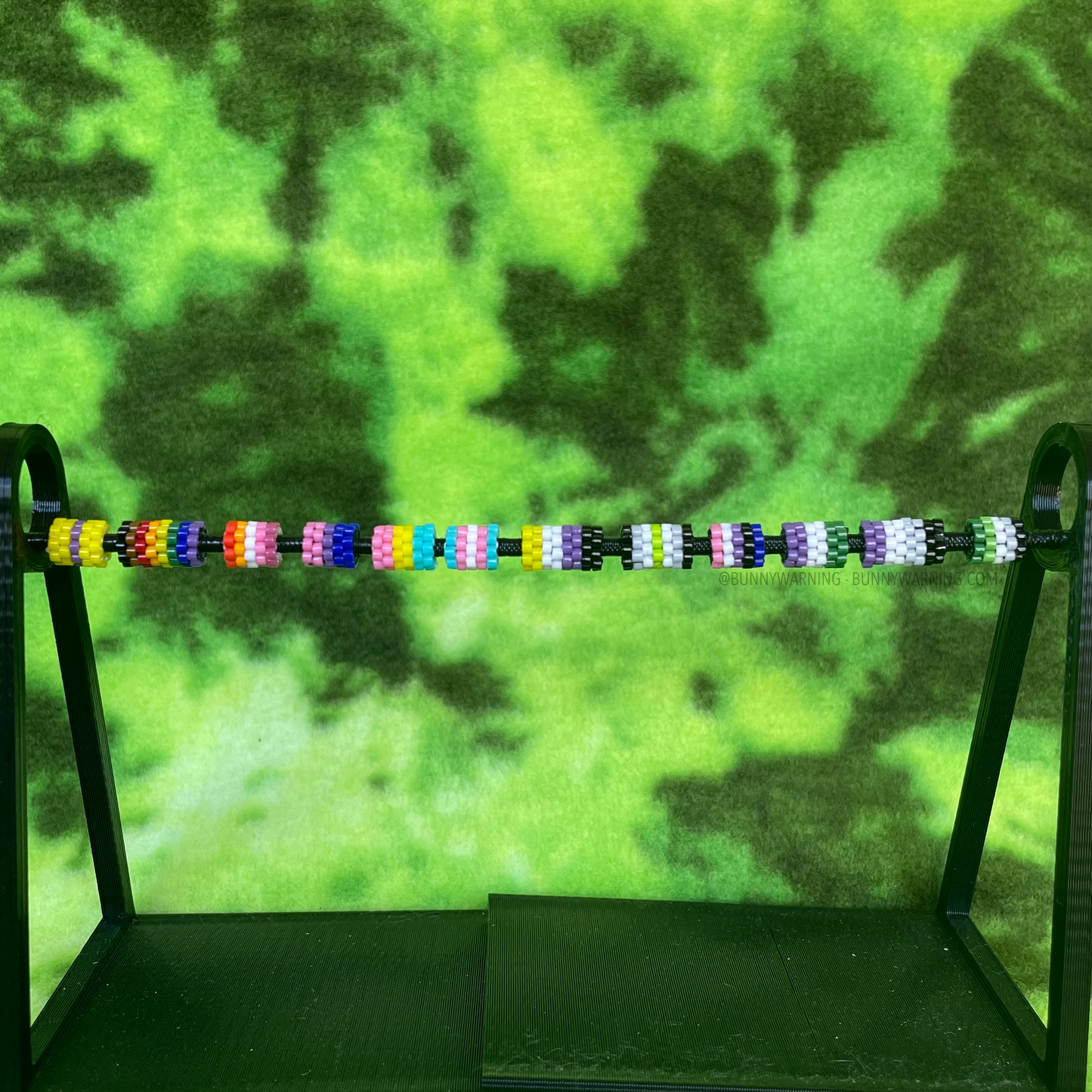 12 beads made of interwoven glass beads made to represent different pride flags. they are displayed horizontally across a black bead display with a light green and dark green tie dye backdrop.